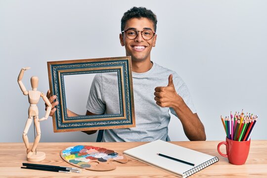 Young handsome african american man painter sitting at art studio holding empty frame smiling happy and positive, thumb up doing excellent and approval sign