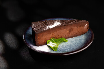 A piece of chocolate cheesecake on a blue plate, decorated with powdered sugar and mint leaves. Black background.