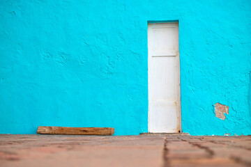 White narrow wooden door in a blue rough painted wall, a wooden log next to a wall, empty space to one side with ample copy space.