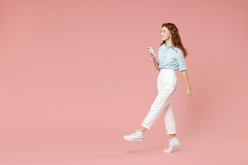 Fototapeta na wymiar Full length side view of young student smiling redhead woman 20s in blue shirt pants using mobile cell phone chatting online in social network walk isolated on pastel pink background studio portrait.