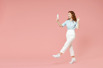 Fototapeta na wymiar Full length side view of young friendly redhead woman in blue shirt pants doing selfie shot on mobile phone talk by video call waving hand greeting isolated on pastel pink background studio portrait.