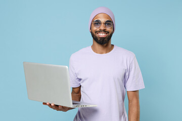 Young smiling unshaven freelancer black african man 20s in violet t-shirt hat glasses hold using...