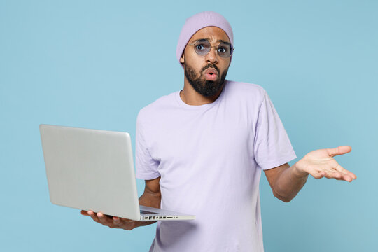 Young confused freelancer black african man in violet t-shirt hat glasses hold use laptop pc computer browse internet spreading hands oops gesture isolated on pastel blue background studio portrait.