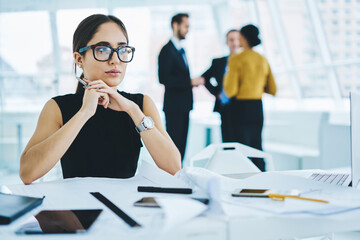 Contemplative business woman in formal wear thinking at table desktop with paper documents and blueprints, thoughtful female executive manager in optical spectacles pondering on idea for project