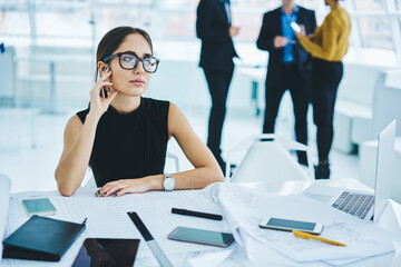 Contemplative business woman in formal wear thinking at table desktop with paper documents and blueprints, thoughtful female executive manager in optical spectacles pondering on idea for project