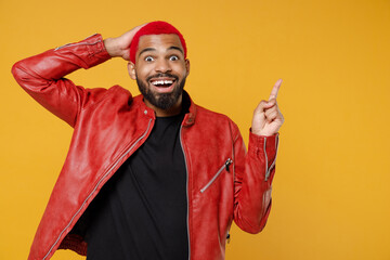 Young african dark-skinned shocked man with funky trendy pink hairdo in red leather jacket point index finger overhead on workspace area hold face isolated on yellow orange background studio portrait.