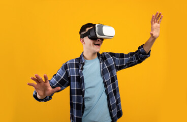 Joyful young guy using virtual reality headset, controlling cyber environment with moving gestures on orange background