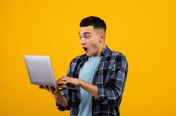 Shocked young guy opening mouth in surprise, looking at laptop screen with unbelievable web content on orange background