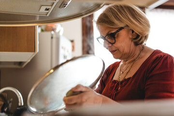 Senior woman cooking lunch in the kitchen - Mature woman in the kitchen lifting the lid of the pot to check the cooking -  Boomer woman busy cooking lunch for the family