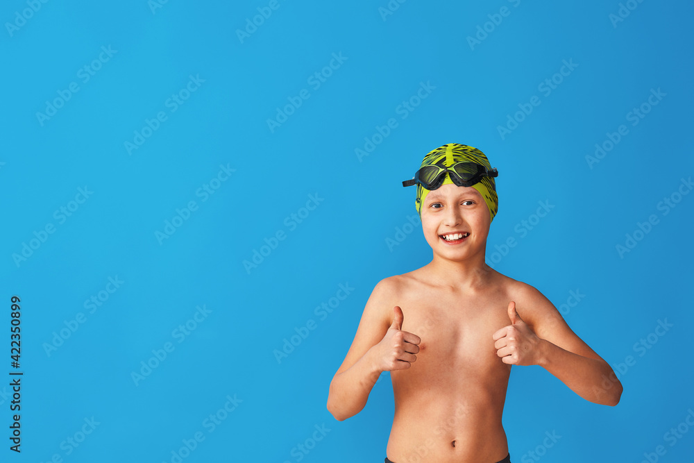 Wall mural small swimmer in a swimming cap, glasses and a bathing suit on a blue background. the child shows thumbs up, sign of approval, advertising. boy is studying at a sports swimming school. Copy space