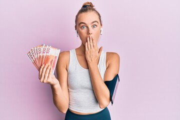 Beautiful blonde sport woman holding 100 norwegian krone banknotes covering mouth with hand, shocked and afraid for mistake. surprised expression