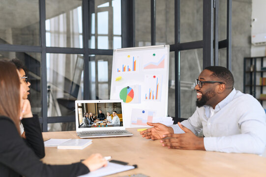 Diverse Work Team Has Virtual Meeting With Outsource Team, Multiracial Colleagues Sit At The Desk In Modern Office Space And Talking Via Video Call With Group On The Laptop Screen, Video Conference