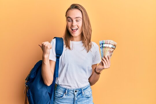 Beautiful blonde woman wearing student backpack and holding canadian dollars pointing thumb up to the side smiling happy with open mouth