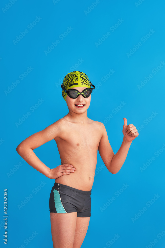 Wall mural small swimmer in a swimming cap, glasses and a bathing suit on a blue background. the child shows thumbs up, sign of approval, advertising. boy is studying at a sports swimming school. Copy space