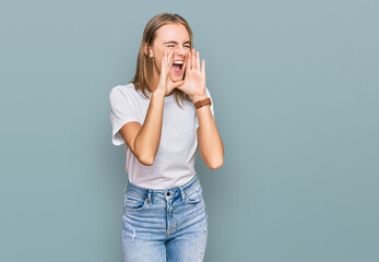 Beautiful young blonde woman wearing casual white t shirt shouting angry out loud with hands over mouth