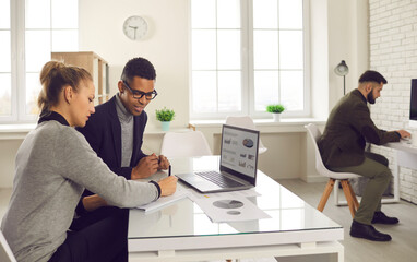 Young diverse people meeting at office desk with laptop computer, working on project, talking, making suggestions and changes, writing business plan, analyzing marketing and sales strategy data charts