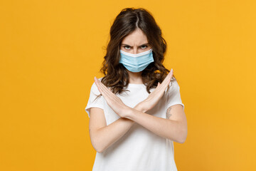 Young serious strict woman in white basic t-shirt in sterile face mask from coronavirus virus covid-19 during pandemic quarantine do stop palm gesture cross hands isolated on yellow orange background.