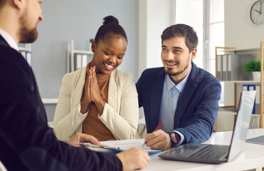 Interracial couple buys real estate. Happy caucasian husband and his dark-skinned wife sign a document while sitting in a bank office. Concept of buying a home or getting mortgage approval.