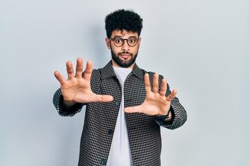 Young arab man with beard wearing glasses afraid and terrified with fear expression stop gesture...