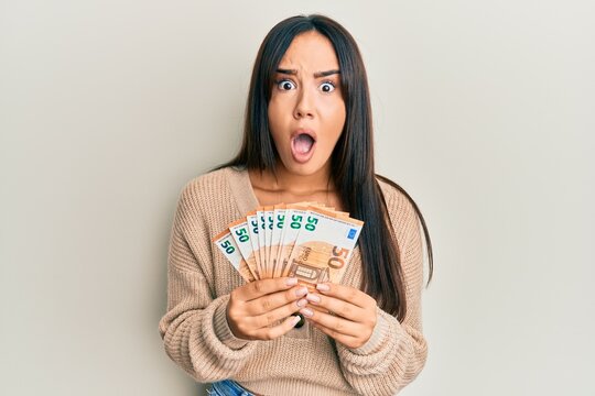 Young beautiful hispanic girl holding bunch of 50 euro banknotes afraid and shocked with surprise and amazed expression, fear and excited face.