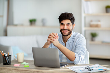 Successful freelancer. Happy arab man sitting at desk with laptop and smiling to camera