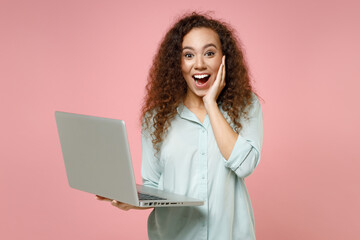 Young black african american shocked surprised fun freelancer curly woman 20s wearing blue shirt holding using laptop pc computer chatting hold face isolated on pastel pink background studio portrait.