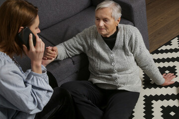Young woman trying to help to grandma on the floor in living room. Woman fell on the floor because...