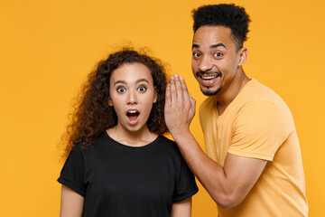 Fototapeta premium Young couple two friends family african shocked man woman in black t-shirt whisper gossip and tell secret behind hand share news with guy look camera isolated on yellow background studio portrait.