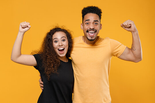 Young couple two american friends family african overjoyed surprised man woman 20s in black yellow t-shirt do winner gesture clench fist celebrating hug isolated on orange background studio portrait