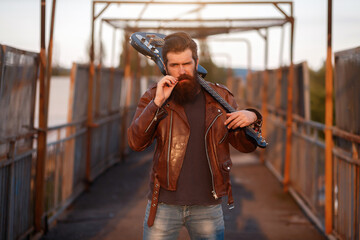 Severe bearded male guitarist with gray hair in a brown leather jacket and blue jeans holds a black electric guitar on his shoulder and straightens his mustache
