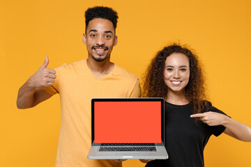 Young couple two friends together family african man woman in black t-shirt point finger on laptop computer blank screen workspace show thumb up gesture isolated on yellow background studio portrait.