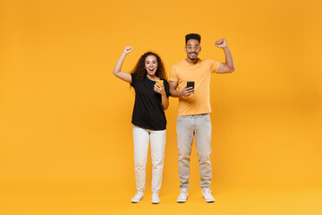 Full length young couple fun two friends together african happy woman man in black tshirt show hold mobile phonedo winner gesture clench fist celebrating isolated on yellow background studio portrait