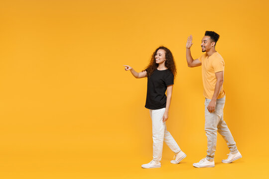 Full Length Side View Young Couple Together Two Friends Family Fun Smiling African Man Woman In Black T-shirt Walking Point Index Finger Wave Hand Greet Isolated On Yellow Background Studio Portrait.