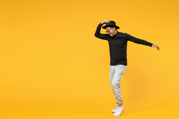 Fototapeta na wymiar Full length young american african man 20s wearing stylish black hat shirt eyeglasses touching hat dancing have fun with outstretched hand isolated on yellow orange color background studio portrait