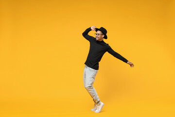 Fototapeta na wymiar Full length young smiling fashionable fun african man 20s wearing stylish black hat shirt eyeglasses stand on toes dancing leaning back dancing isolated on yellow orange background studio portrait