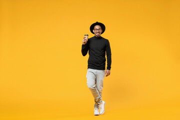 Fototapeta na wymiar Full length young happy smiling fun american african man 20s wear stylish black shirt hat eyeglasses drinking coffee hot tea in paper cup isolated on yellow orange color background studio portrait