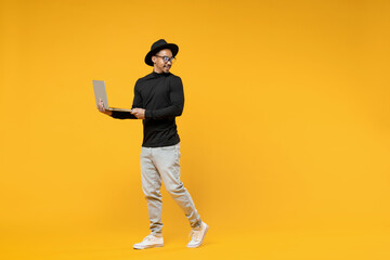 Full length young freelancer smiling african man in stylish black shirt hat eyeglasses using laptop pc computer chat in social network look aside isolated on yellow orange background studio portrait