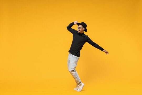 Full Length Young Smiling Fashionable Fun African Man 20s Wearing Stylish Black Hat Shirt Eyeglasses Stand On Toes Dancing Leaning Back Dancing Isolated On Yellow Orange Background Studio Portrait