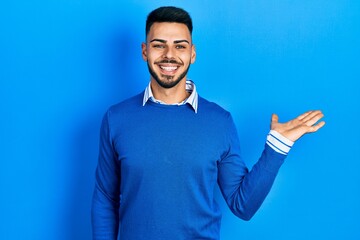 Young hispanic man with beard wearing casual blue sweater smiling cheerful presenting and pointing with palm of hand looking at the camera.