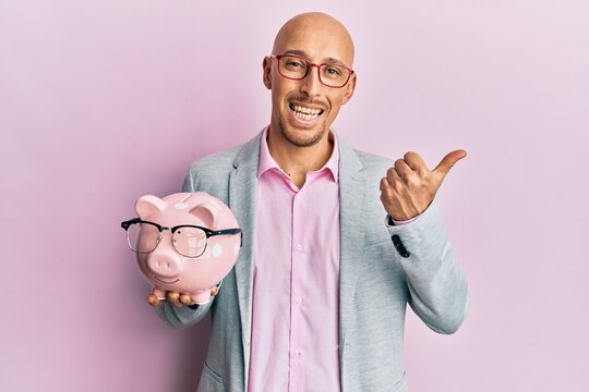 Bald man with beard holding piggy bank with glasses pointing thumb up to the side smiling happy with open mouth