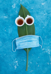 Two white cups with tea and a mask standing on a green leaf with blue background