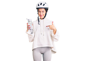 Beautiful brunette young woman wearing bike helmet and holding water bottle pointing finger to one self smiling happy and proud