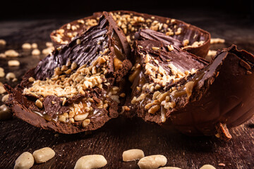Stuffed chocolate easter egg with peanut and caramel on a wooden table with lots of peanuts around. Cracked. Close up.