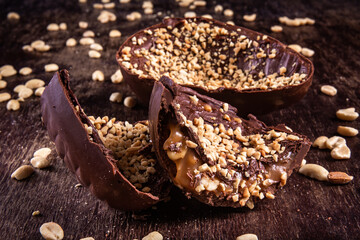 Stuffed chocolate easter egg with peanut and caramel on a wooden table with lots of peanuts around. Cracked. Close up.