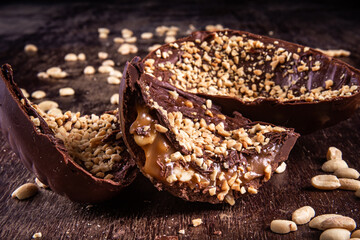 Stuffed chocolate easter egg with peanut and caramel on a wooden table with lots of peanuts around. Cracked. Close up.