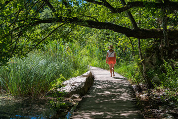 Obraz premium Woman in sunny hat walking on wooden pavement in forest with hidg grass and rushes.