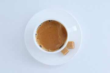 White cup of coffee with froth and cane sugar cubes on a saucer on white background.