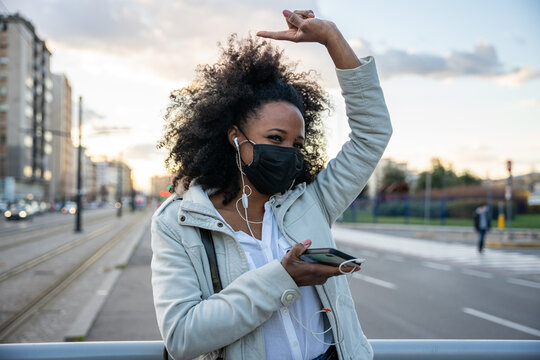 Portrait of a young woman in the evening at sunset in the city wearing protective face mask during global pandemic from Covid-19 Coronavirus dancing listening to music with earphones from smartphone