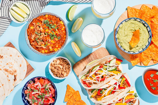 Mexican food table with traditional dishes. Chili con carne, tacos, tomato salsa, corn chips with guacamole. Mexican feast in hard light on blue color background, top view