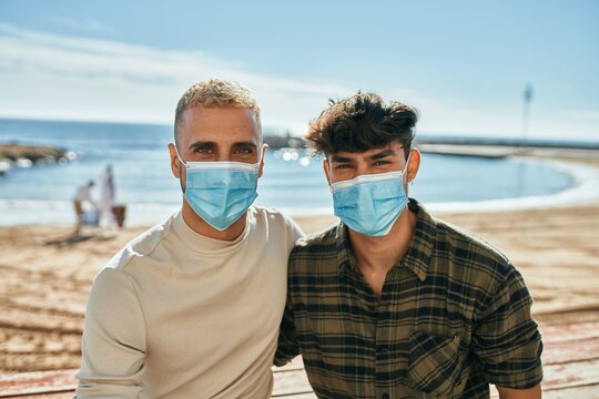 Young gay couple wearing medical mask sitting on the bench at the beach.